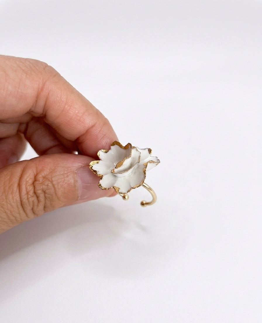 A hand holding a white flower shaped ceramic ring with gold edges