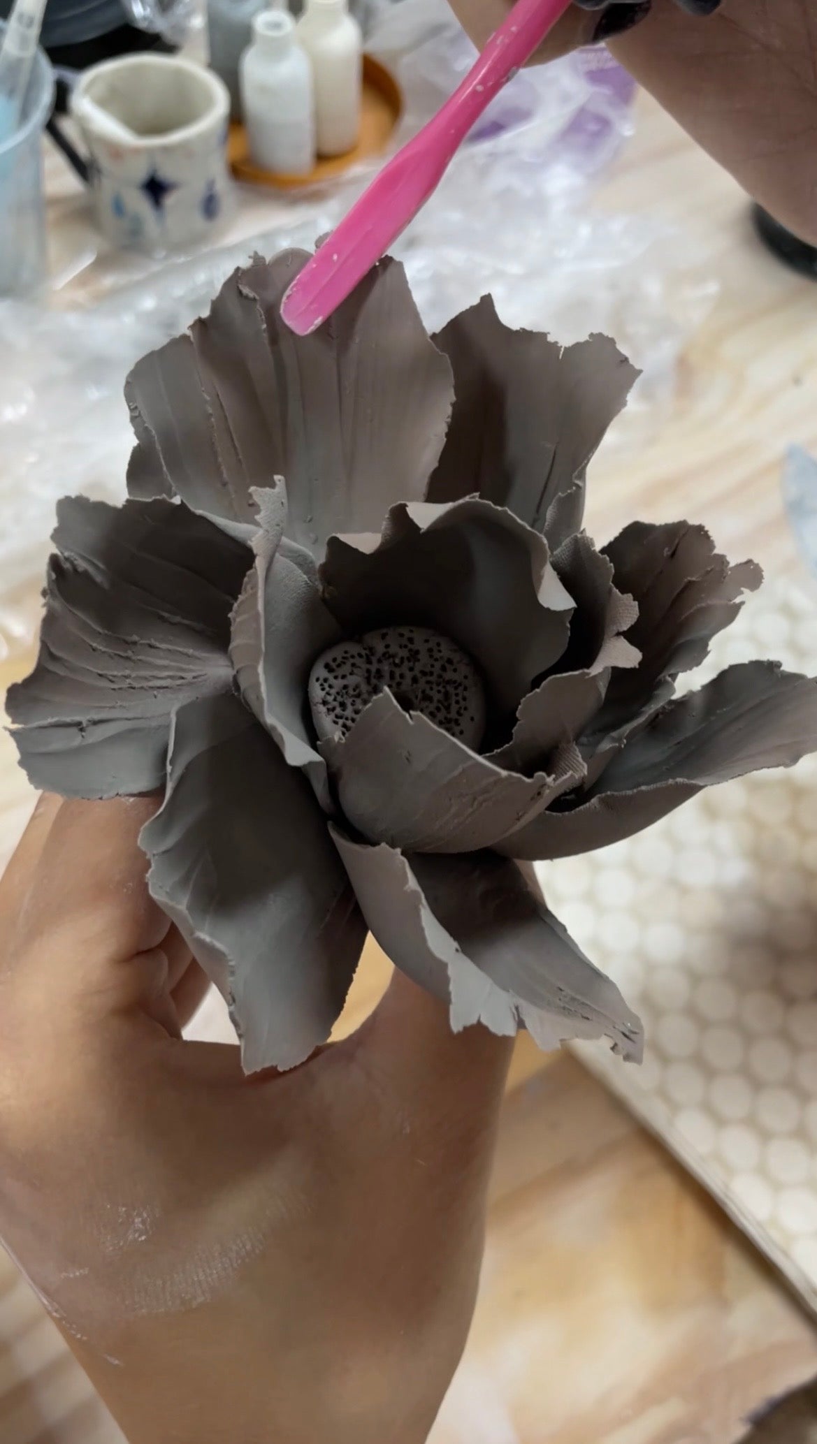 Hands sculpting a grey clay flower with textured petals using a pink tool